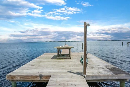 A wooden dock extends over calm water, with a table at the end and a cloudy blue sky above.
