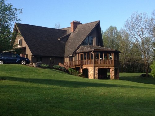 A large, modern house with a stone porch, surrounded by green grass and trees under a clear blue sky.