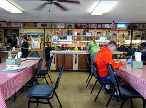 A cozy diner interior with checkered tablecloths, patrons eating, and a counter with food preparation in the background.