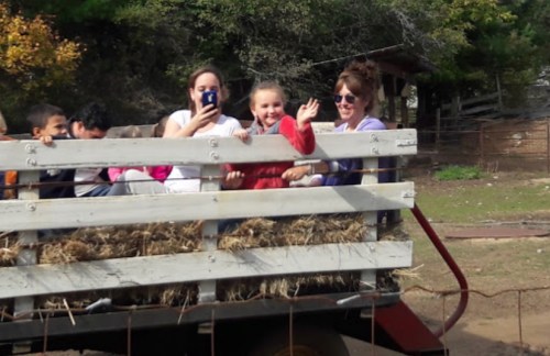 A group of children and adults smiling and waving from a hayride on a sunny day.