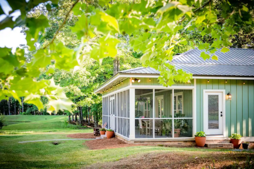 A charming green house with a screened porch, surrounded by lush trees and a grassy yard.