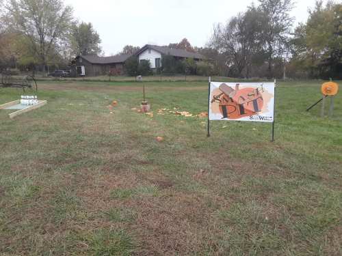 A grassy field with a sign for a pumpkin-smashing event, scattered pumpkins, and a wooden structure in the background.