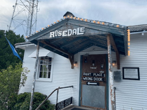 A rustic building with a sign reading "Rosedale" and "Right Place, Wrong Door" above the entrance, decorated with lights.