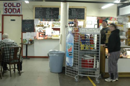 A small café interior with a menu board, snacks on display, and customers at the counter. A "COLD SODA" sign is visible.