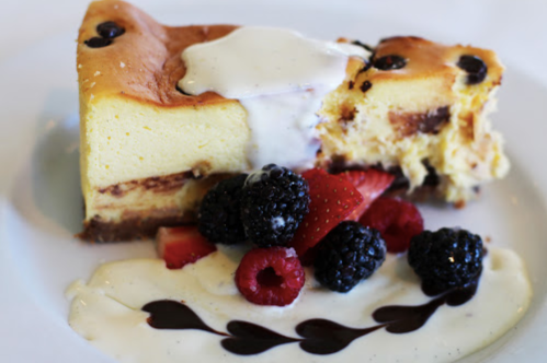 A slice of cheesecake topped with cream, surrounded by fresh berries and a drizzle of chocolate sauce on a white plate.