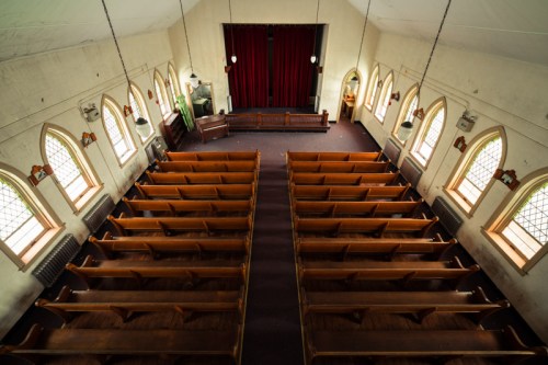 Interior of a church with wooden pews arranged in rows and a red curtain at the front, illuminated by natural light.