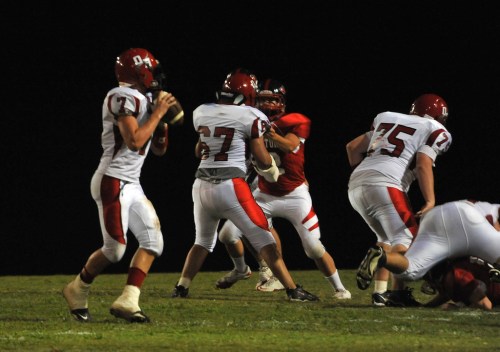 A football game scene with players in red and white uniforms, one quarterback preparing to throw while others engage.