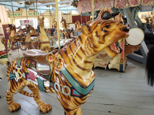 A colorful, vintage carousel tiger with a saddle, set in an amusement park with other carousel animals in the background.