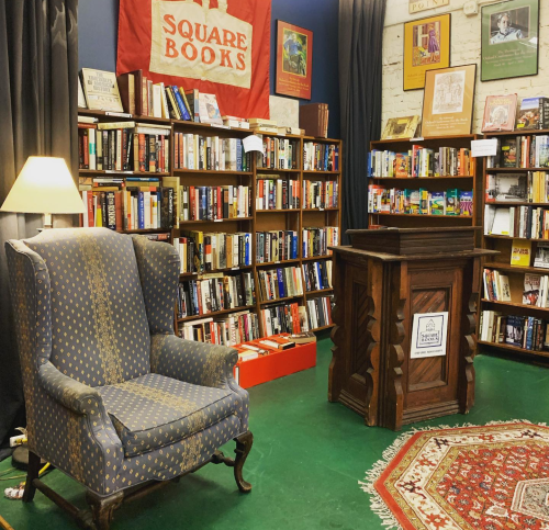 Cozy bookstore corner with a blue armchair, wooden podium, and shelves filled with books against a colorful backdrop.