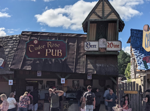 A bustling pub named "Tudor Rose" with a wooden exterior, people gathered outside, and signs for beer and wine.