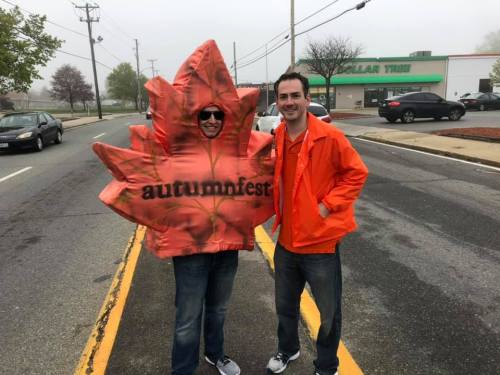 Two people stand on a foggy street, one wearing a large inflatable leaf costume labeled "autumnfest," the other in an orange jacket.