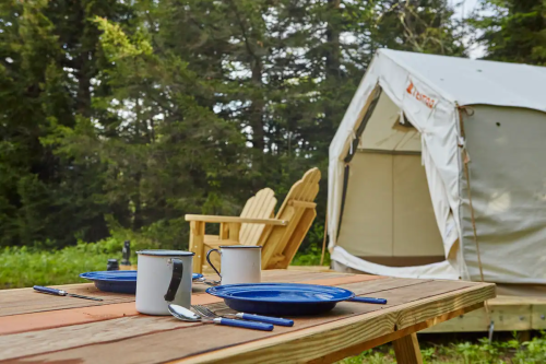 A cozy campsite with a wooden table set for two, featuring blue plates and mugs, beside a glamping tent in a forest.