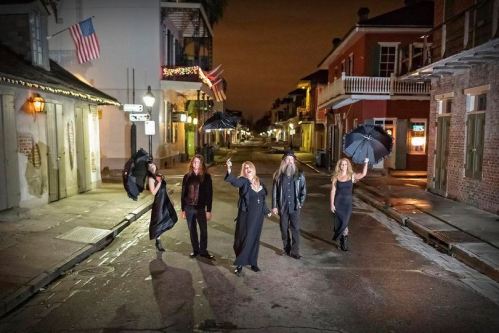 Five people in black attire holding umbrellas stand in a dimly lit street, with historic buildings and flags in the background.