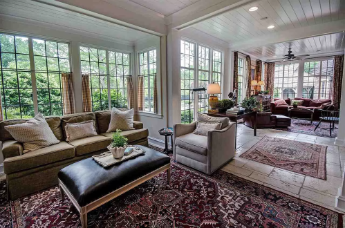 Bright sunroom with large windows, cozy seating, and a patterned rug, featuring plants and warm lighting.