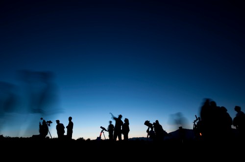 Silhouettes of people with telescopes against a twilight sky, observing stars and celestial objects.