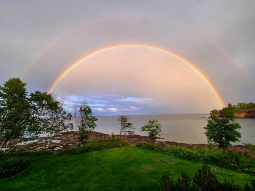 A vibrant double rainbow arcs over a calm lake, framed by lush greenery and rocky shoreline.