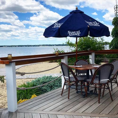 A beachside patio with a table and chairs under a blue umbrella, overlooking a sandy shore and calm water.