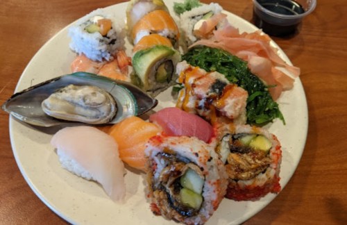 A plate of assorted sushi, including rolls, sashimi, and seaweed salad, with soy sauce and pickled ginger on the side.