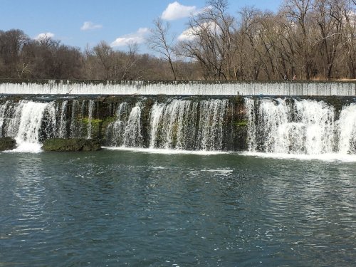 A serene waterfall cascading over rocks into a calm river, surrounded by bare trees and a blue sky with fluffy clouds.