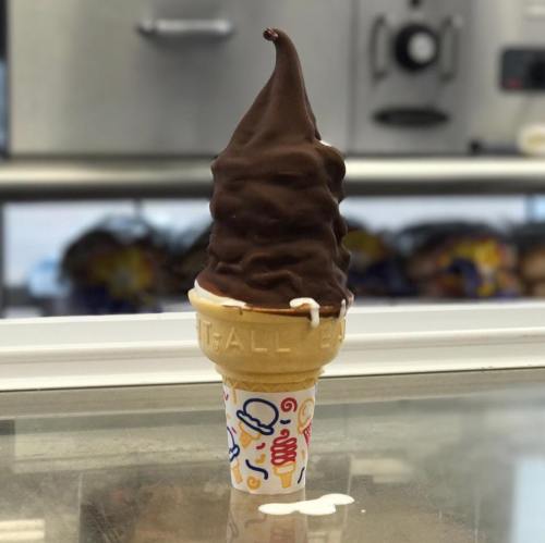 A chocolate soft serve ice cream cone with a colorful patterned cup, sitting on a counter with a blurred background.