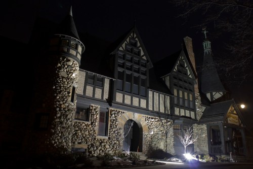 A large, ornate stone mansion illuminated at night, featuring gothic architecture and intricate details.