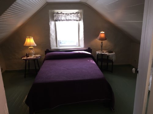 Cozy attic bedroom with a purple bedspread, two lamps, and a window letting in natural light.