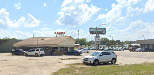 A small restaurant building with a sign, surrounded by parked cars and a billboard under a partly cloudy sky.