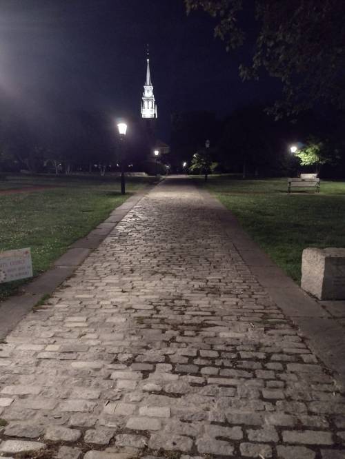 A cobblestone path at night, lined with lampposts, leading to a church steeple illuminated in the distance.