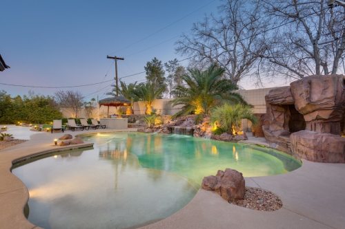 A serene backyard pool surrounded by palm trees, rocks, and soft lighting, creating a tranquil evening atmosphere.