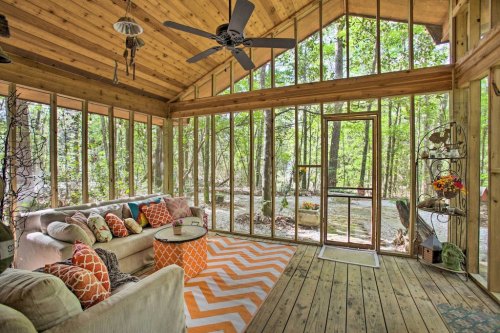 Cozy sunroom with large windows, wooden ceiling, comfortable seating, and a vibrant orange chevron rug. Surrounded by trees.