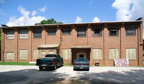 Abandoned brick building with boarded windows, two parked cars in front, under a blue sky with scattered clouds.