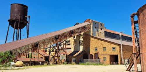 Abandoned industrial building with a sloped roof, water tower, and rusty structures under a clear blue sky.