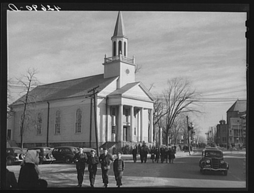 A group of people walking past a church on a street, with vintage cars parked nearby and trees in the background.