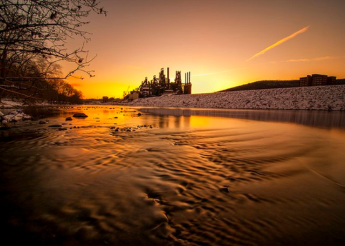 Sunset over a river with industrial buildings in the background, reflecting warm colors on the water's surface.