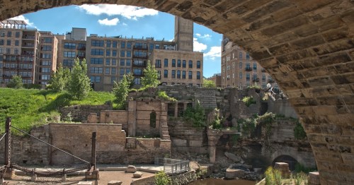 View of historic ruins and greenery under a stone arch, with modern buildings in the background against a blue sky.