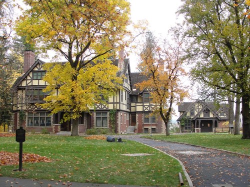A large, historic house with a Tudor-style design, surrounded by autumn trees and fallen leaves.