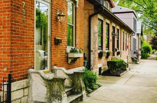 A charming street view of brick houses with flower boxes and stone steps, lined with greenery and a clear sidewalk.