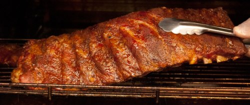 Close-up of a perfectly cooked rack of ribs being lifted with tongs from a smoker, glistening with barbecue sauce.