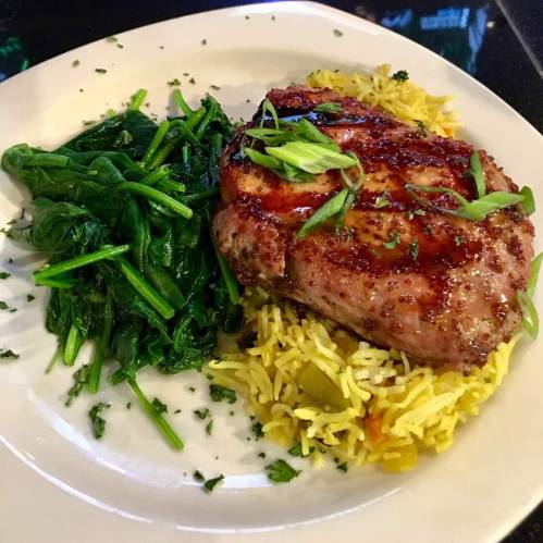 A plate featuring grilled pork chop on yellow rice, served with sautéed spinach and garnished with green onions.
