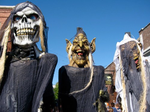 Three large, spooky puppets with skull and goblin faces, dressed in tattered black clothing against a clear blue sky.