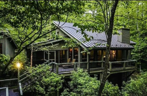 A modern cabin nestled in lush greenery, featuring large windows and a wraparound deck.