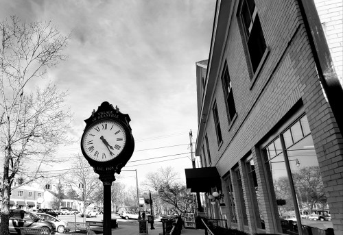 A vintage clock stands in front of a brick building, with trees and a cloudy sky in the background.