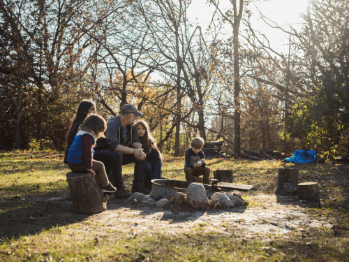 A family sits around a campfire in a wooded area, enjoying time together on a sunny day.