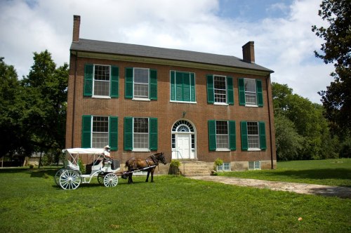 A historic brick house with green shutters, featuring a horse-drawn carriage in front on a grassy lawn.