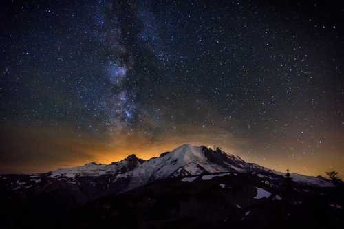 A stunning night sky filled with stars above a snow-capped mountain, illuminated by a soft glow from the horizon.