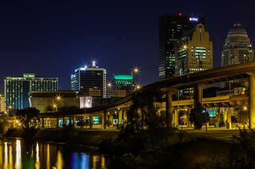 Night view of a city skyline with illuminated buildings and a highway overpass reflecting in the water below.