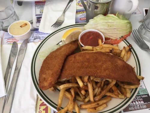 A plate of fried fish fillets served with French fries, a lemon wedge, lettuce, tomato, and a small cup of sauce.