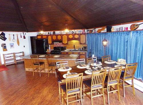 A spacious dining area with a large wooden table, chairs, and a kitchen in the background, featuring blue curtains.