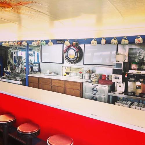A retro diner interior featuring a counter, coffee machines, and hanging mugs, with red and blue decor.