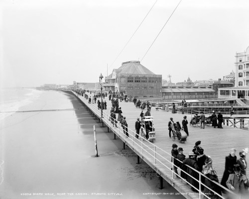 Historic black-and-white photo of a busy boardwalk in Atlantic City, with people walking near the casino.